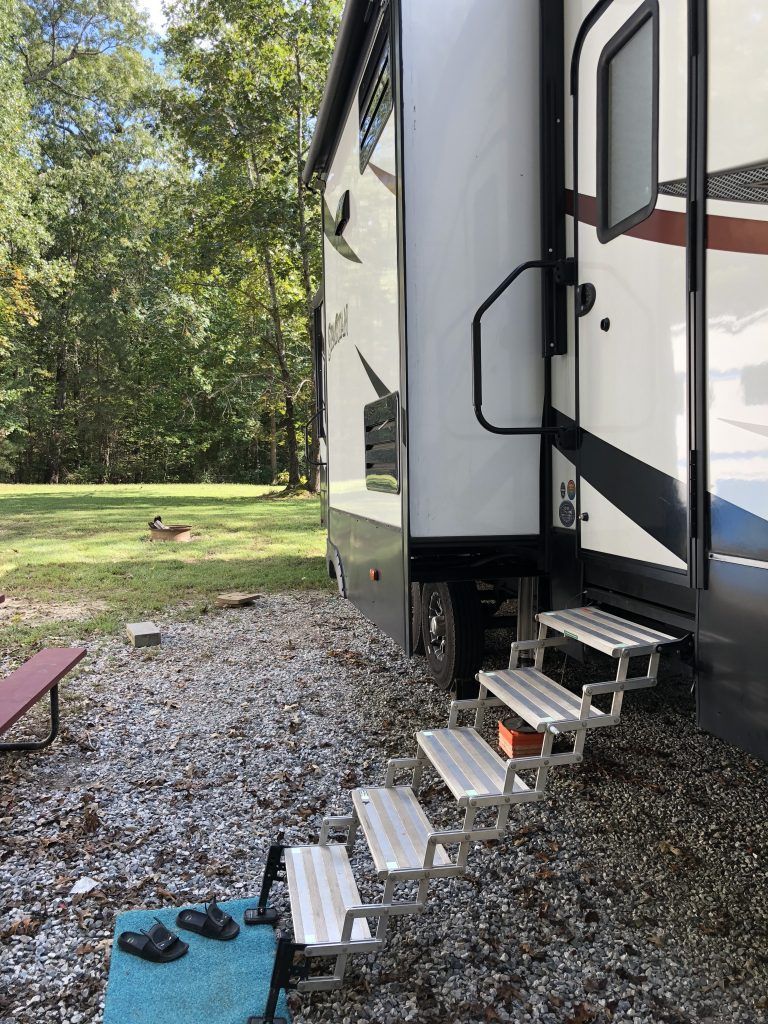 A rv with stairs leading up to it is parked in a gravel lot.