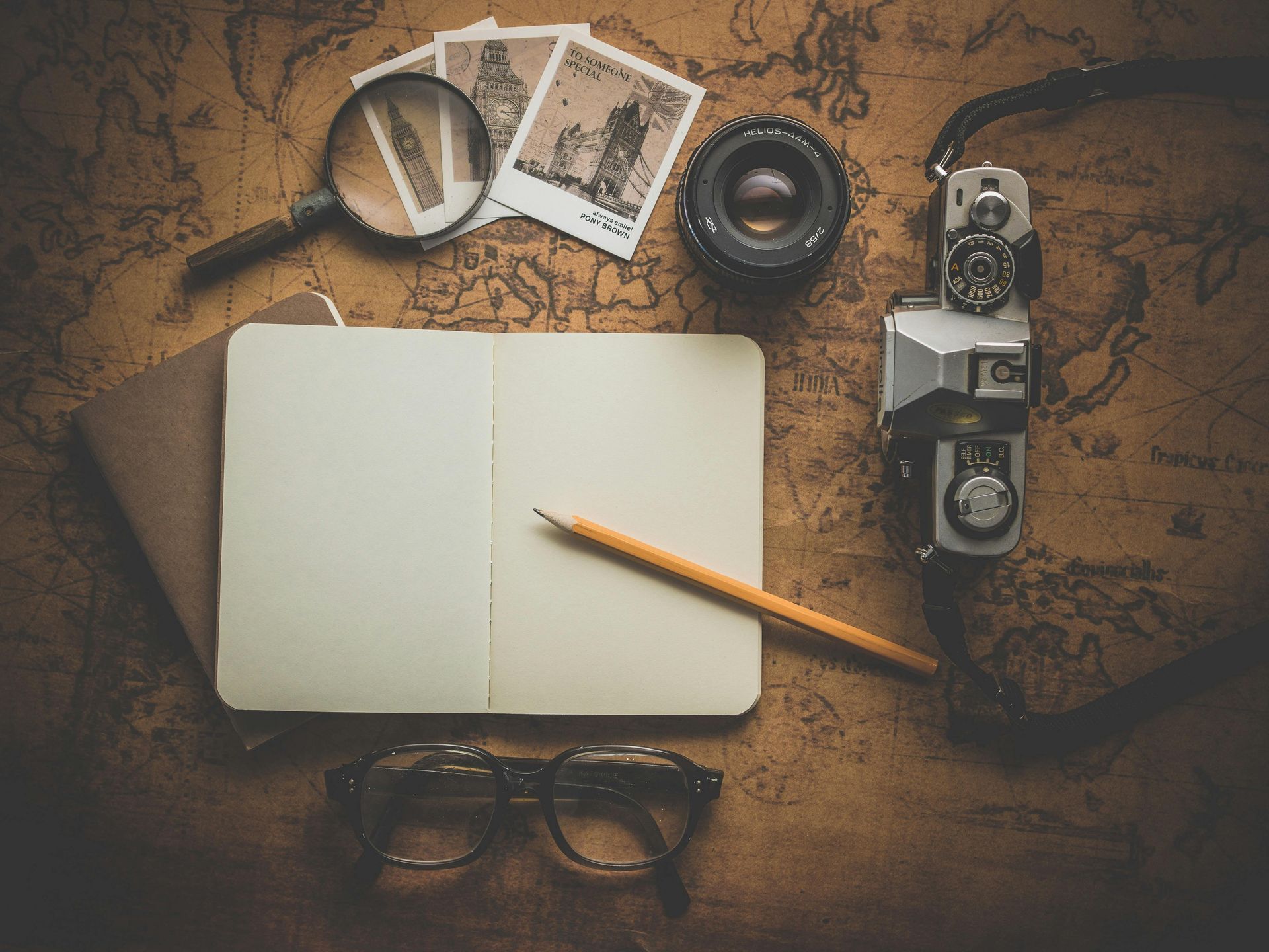 A notebook , camera , magnifying glass , glasses and a pencil are on a map.