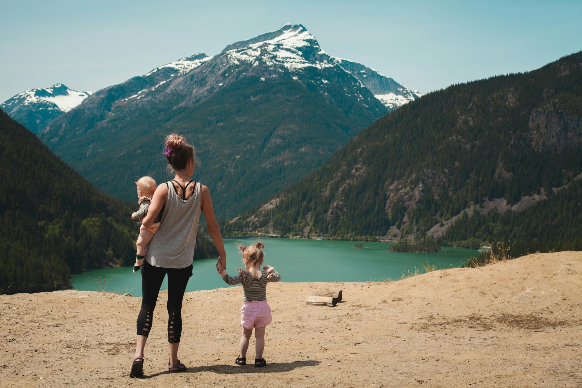 A woman is holding a baby and a little girl while standing on top of a hill overlooking a lake.
