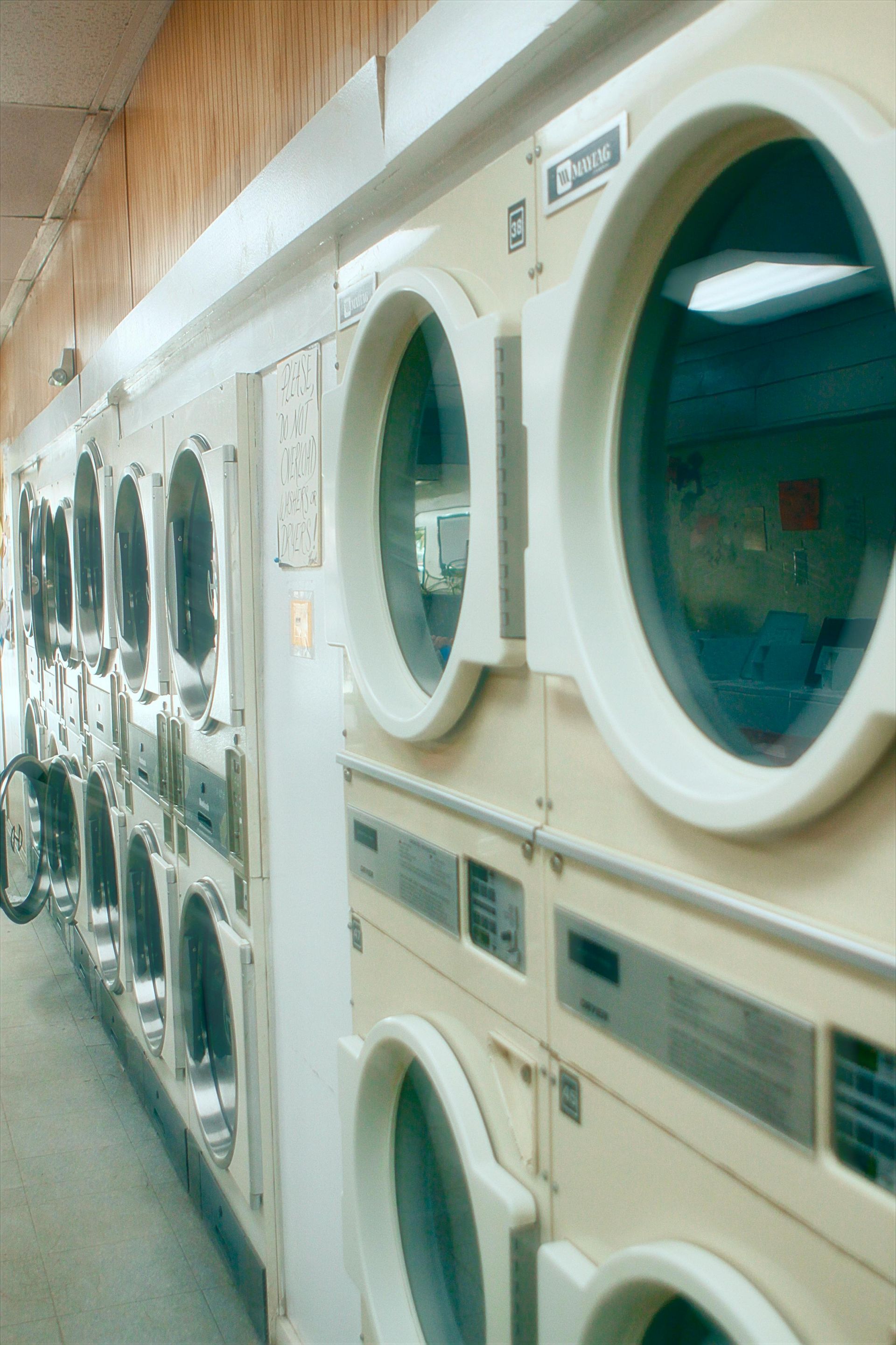 A long row of stacked, light-colored commercial laundry dryers with circular glass doors in a laundromat.