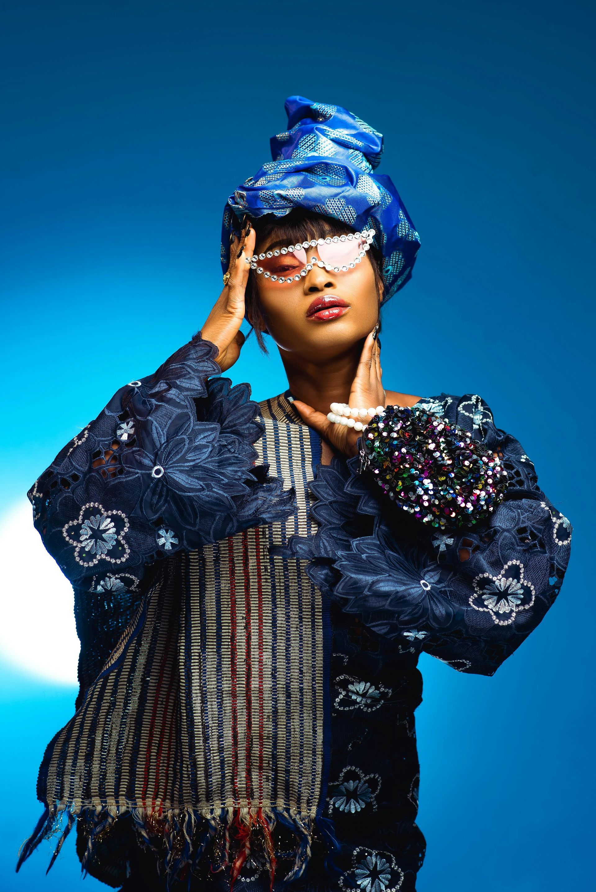 A woman wearing a head scarf and sunglasses is standing in front of a blue background.