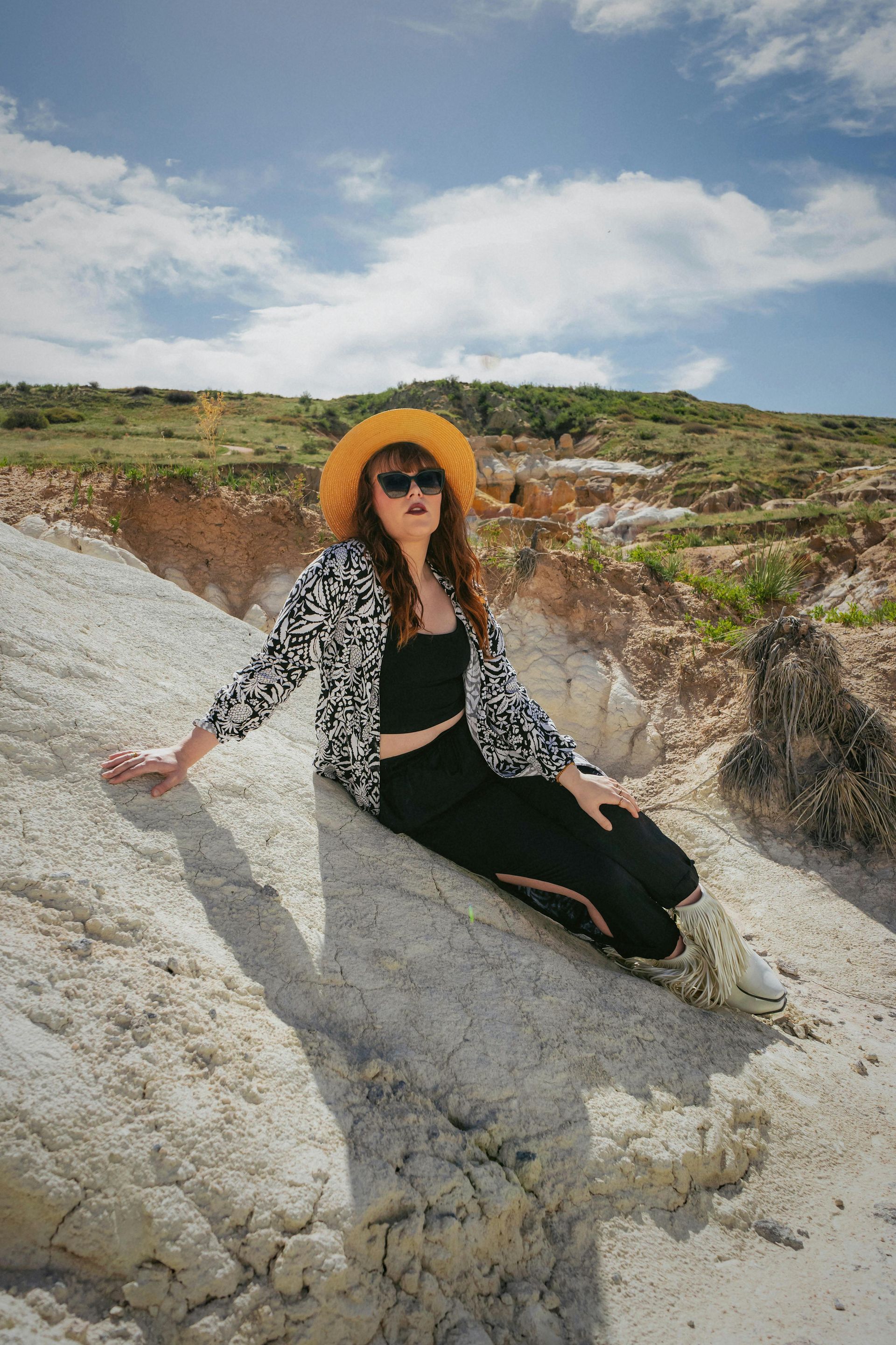 A woman is sitting on a rock wearing a hat and sunglasses.