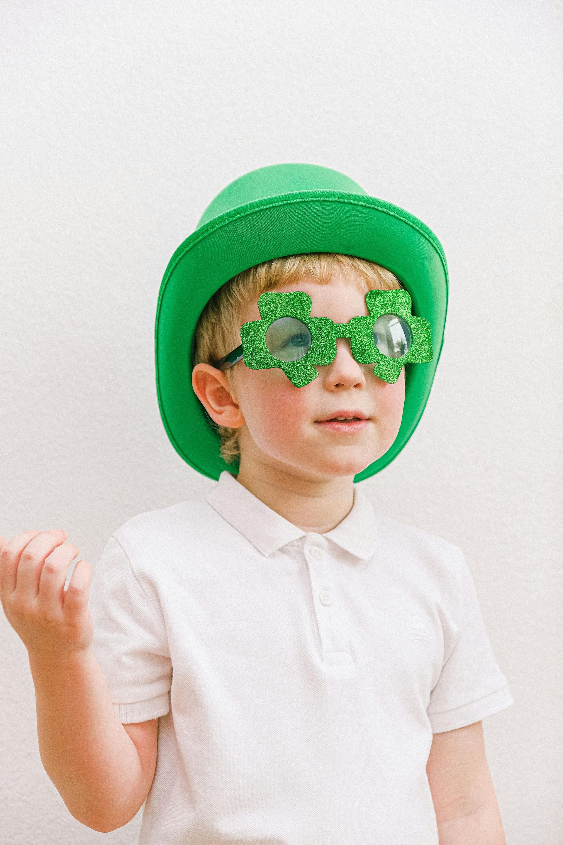 Boy in green hat and shamrock glasses, raising a hand. White background.