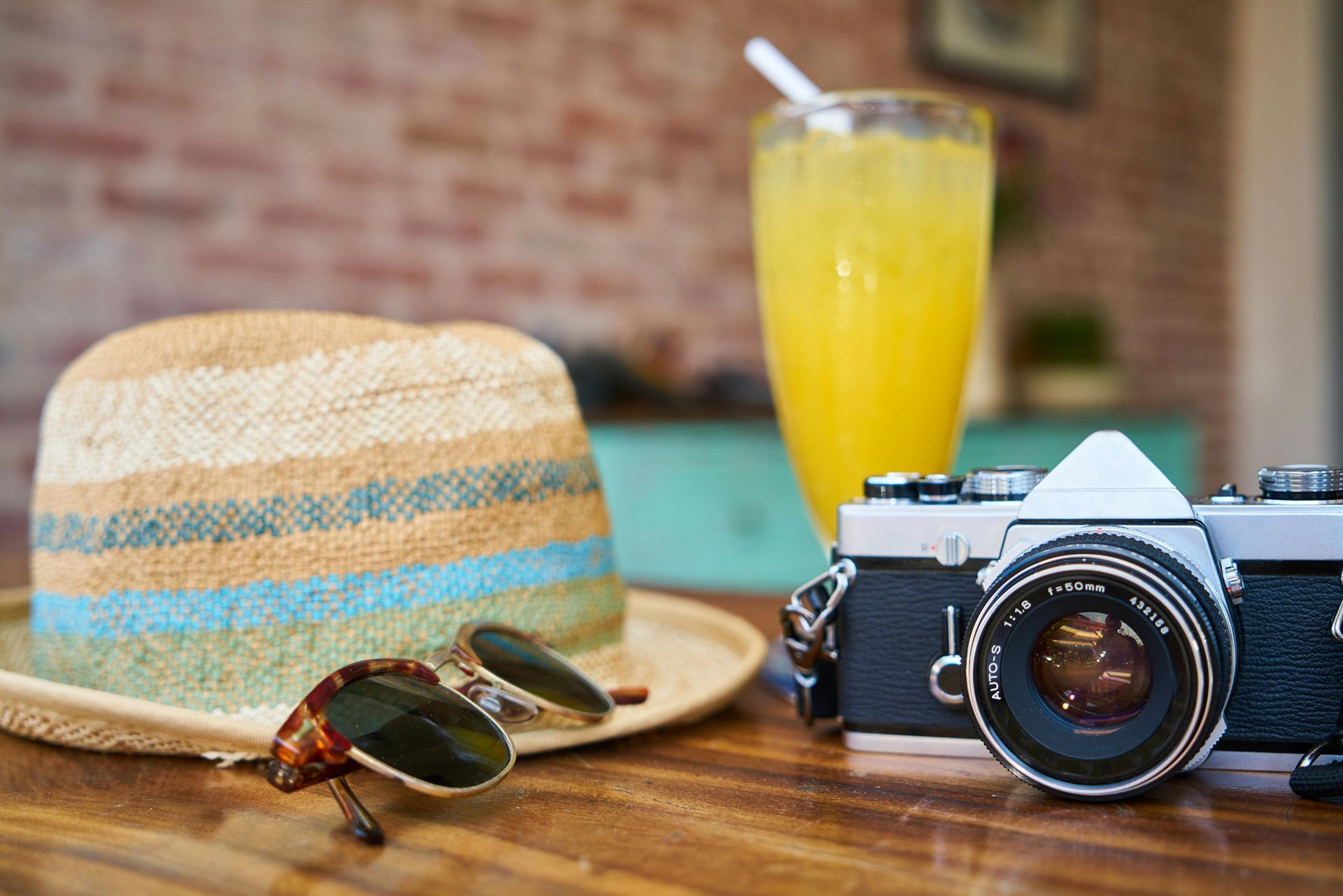 A hat , sunglasses , a camera , and a glass of orange juice are on a wooden table.