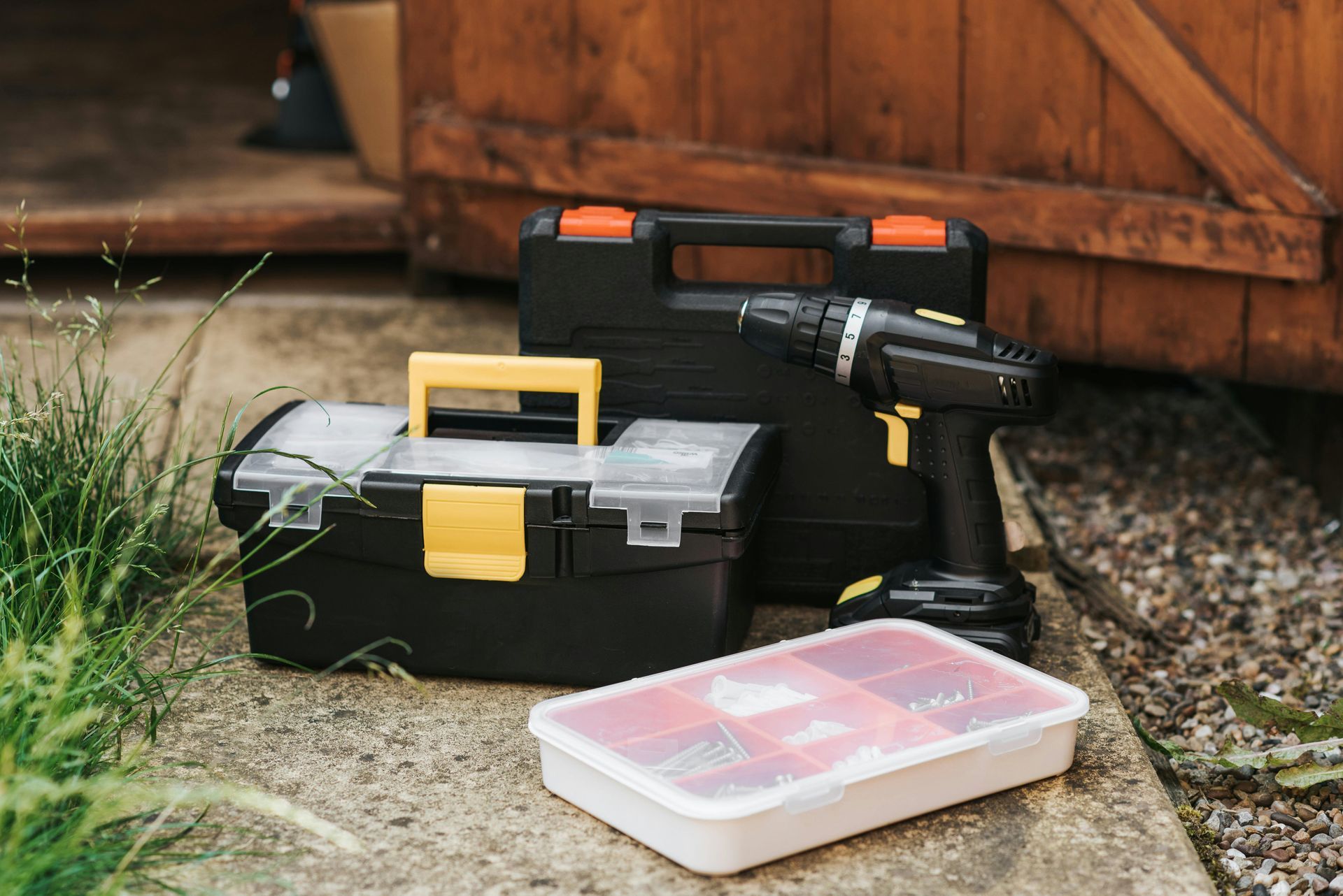 A drill and a toolbox are sitting on the ground next to a wooden shed.