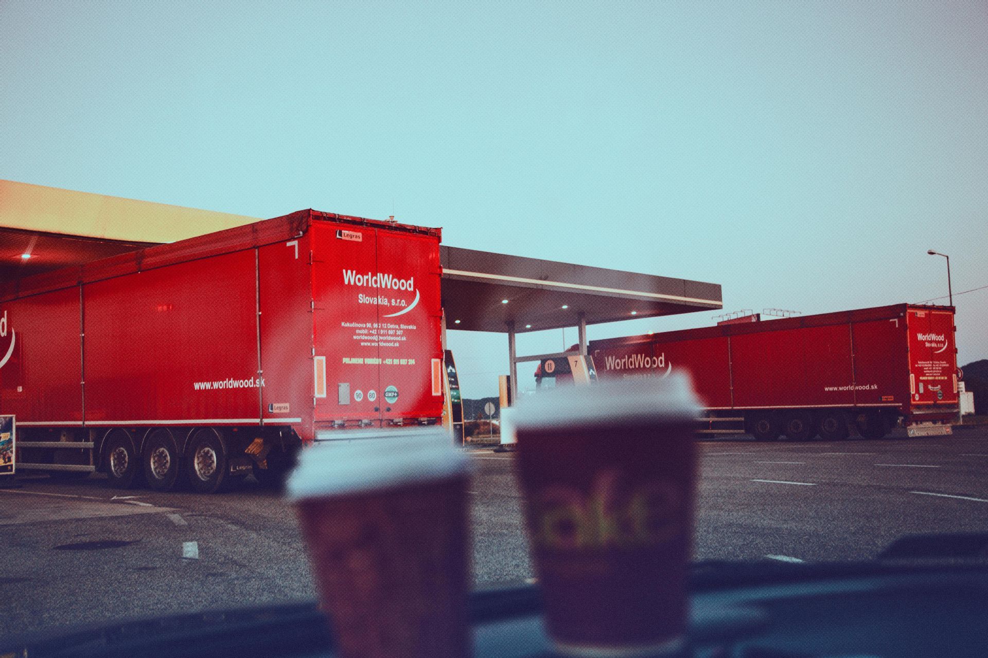 Two coffee cups sit in the foreground with two red articulated lorries parked at a service station behind them.