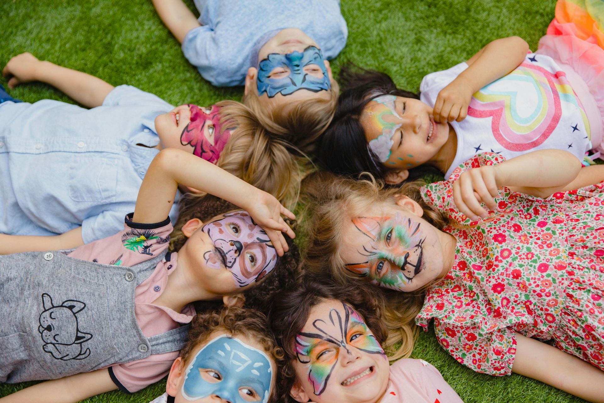 A group of children with their faces painted are laying in a circle on the grass.