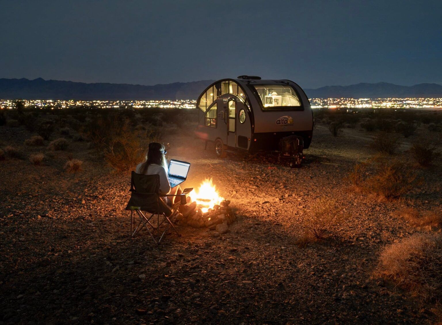 A person is sitting at a campfire with a laptop in front of a trailer.