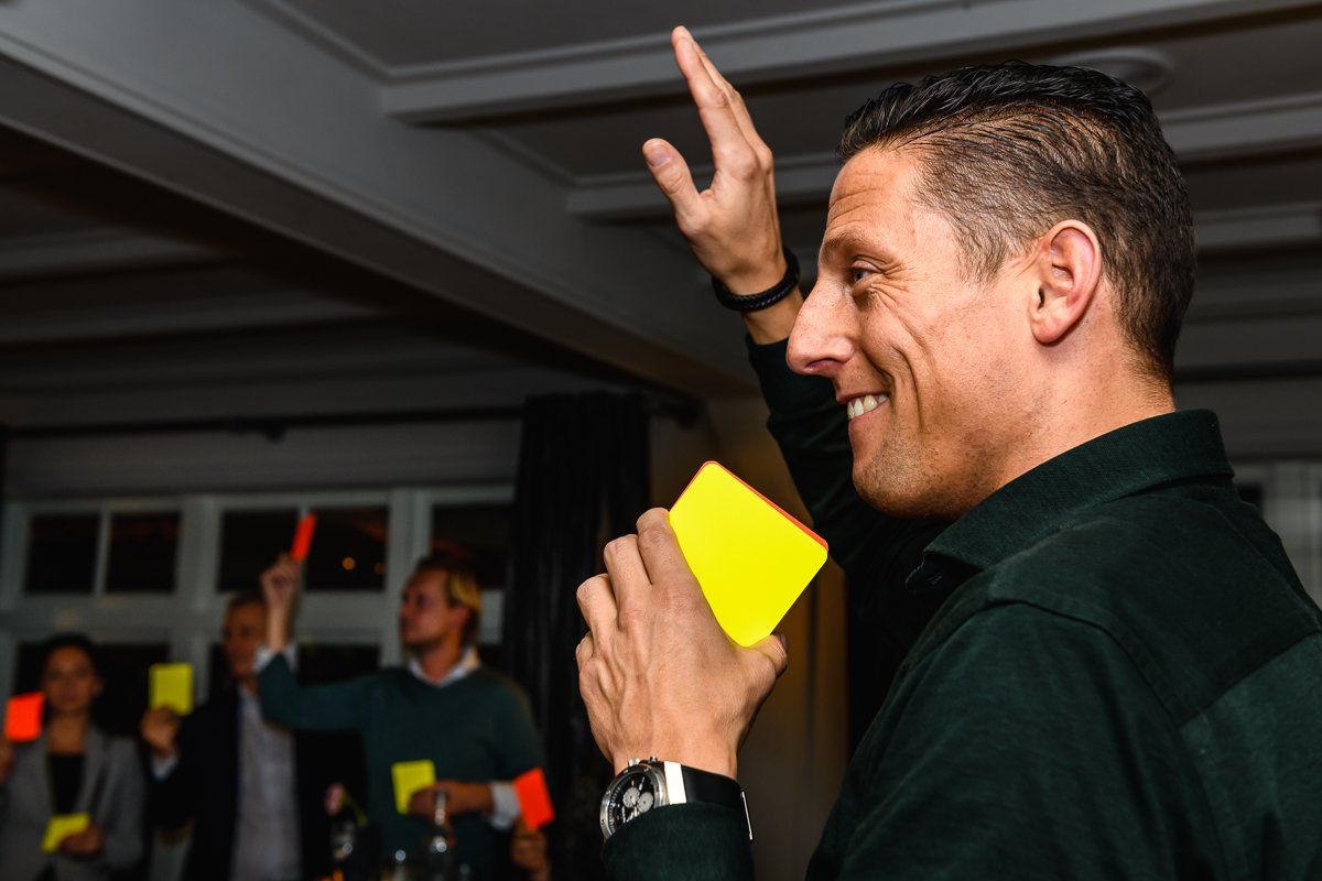 Een man houdt een gele kaart omhoog en lacht. Anderen houden kaarten omhoog in een ruimte met warme verlichting.