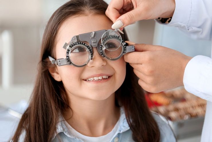 Una ragazza con gli occhiali sorride durante un esame della vista, con una tabella oculistica sullo sfondo.