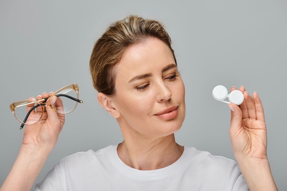 Donna che tiene in mano occhiali e astuccio per lenti a contatto, con aria contemplativa su uno sfondo grigio.