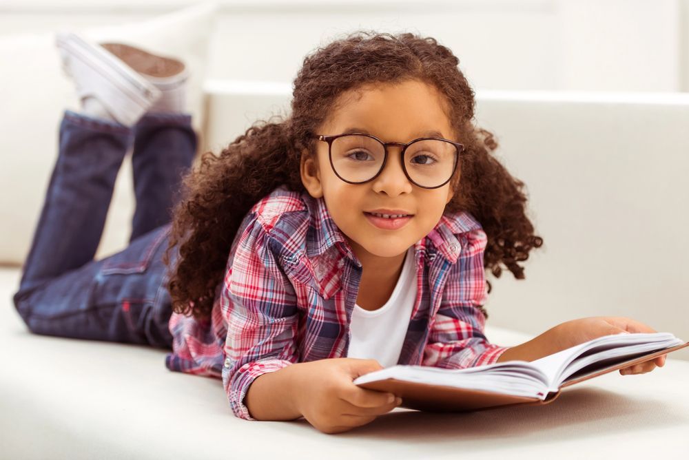 Ragazza con gli occhiali, che legge un libro sdraiata sul divano, indossando una camicia a quadri e jeans.