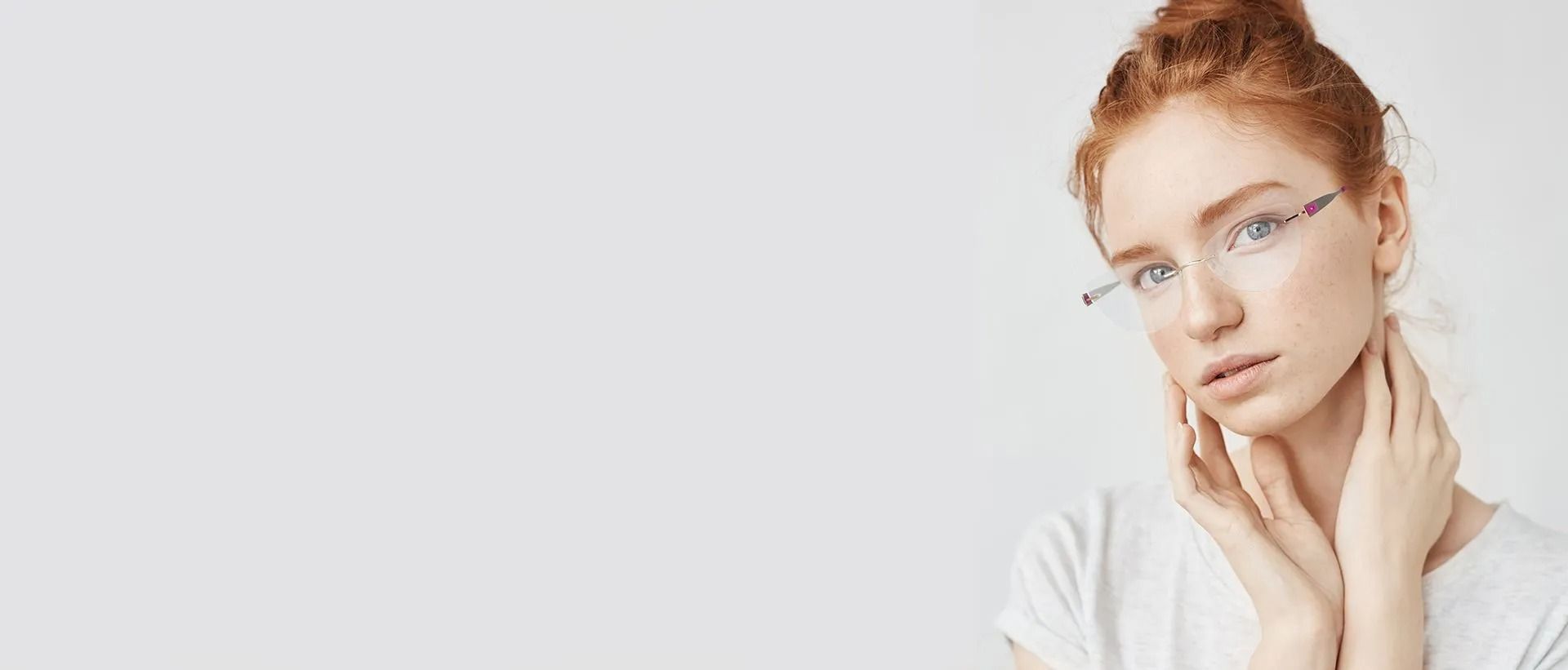 Donna con i capelli rossi e gli occhiali, che si tocca il collo, camicia bianca, su sfondo bianco.