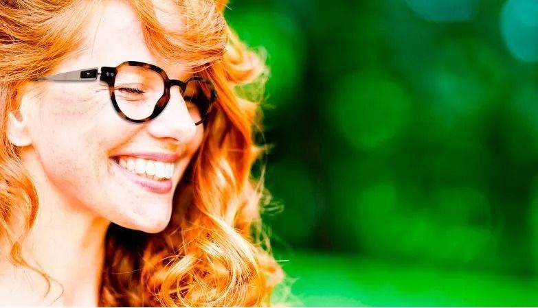 Una donna con i capelli rossi e gli occhiali sorride ampiamente su uno sfondo verde.