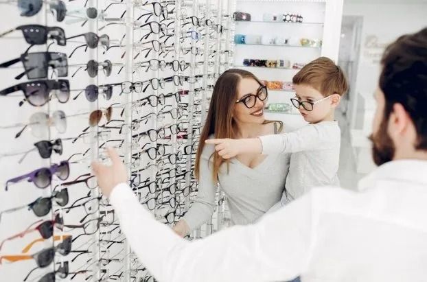 Famiglia che sceglie gli occhiali in un negozio di ottica. La madre indica, il figlio indossa gli occhiali, l'ottico aiuta.