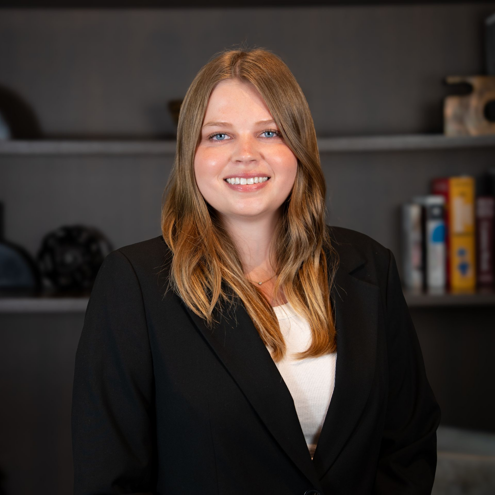 Smiling woman in a black blazer standing in front of a bookshelf in an office setting