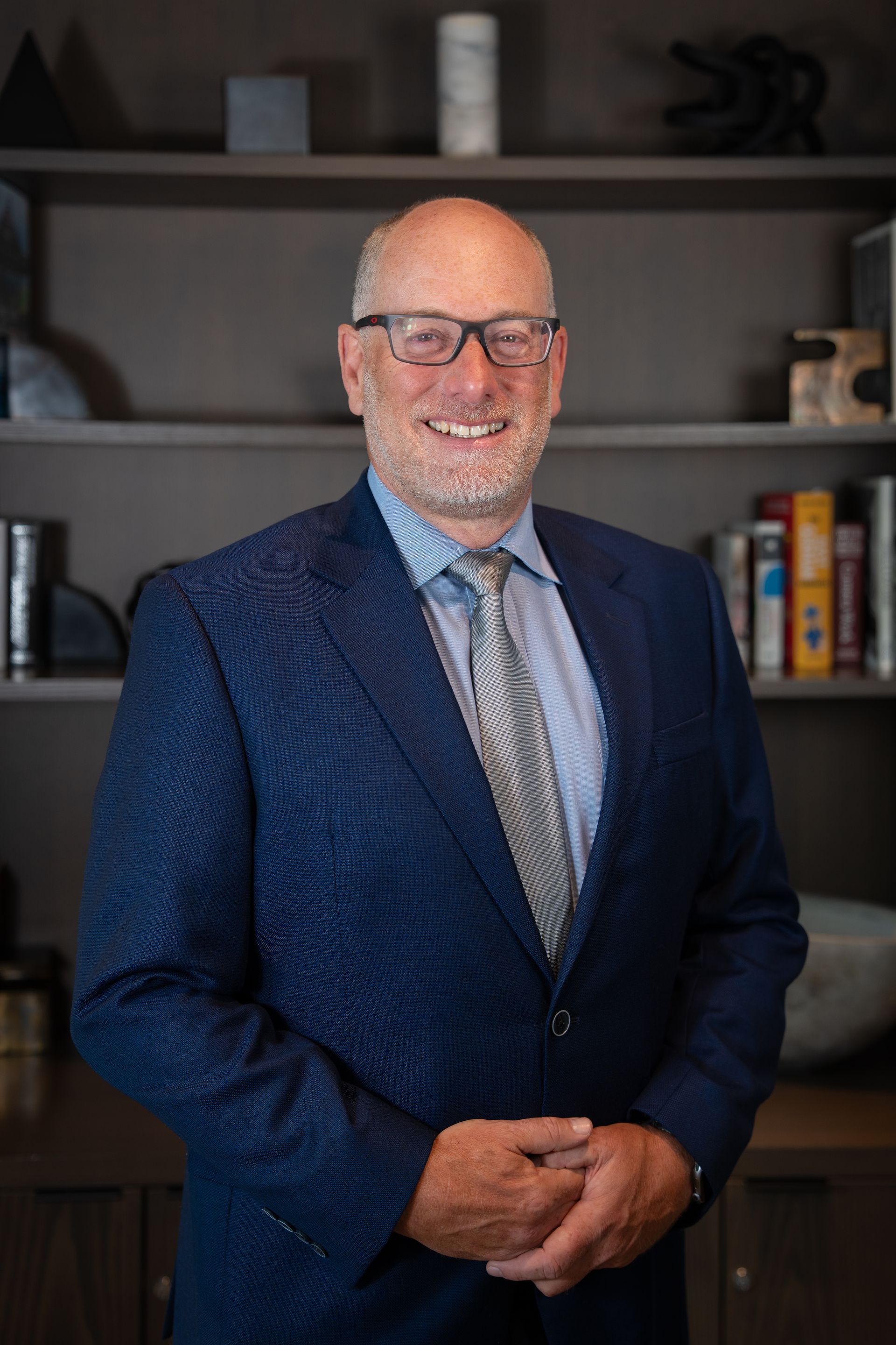 Man in a blue suit and tie standing indoors in front of shelves, smiling at the camera