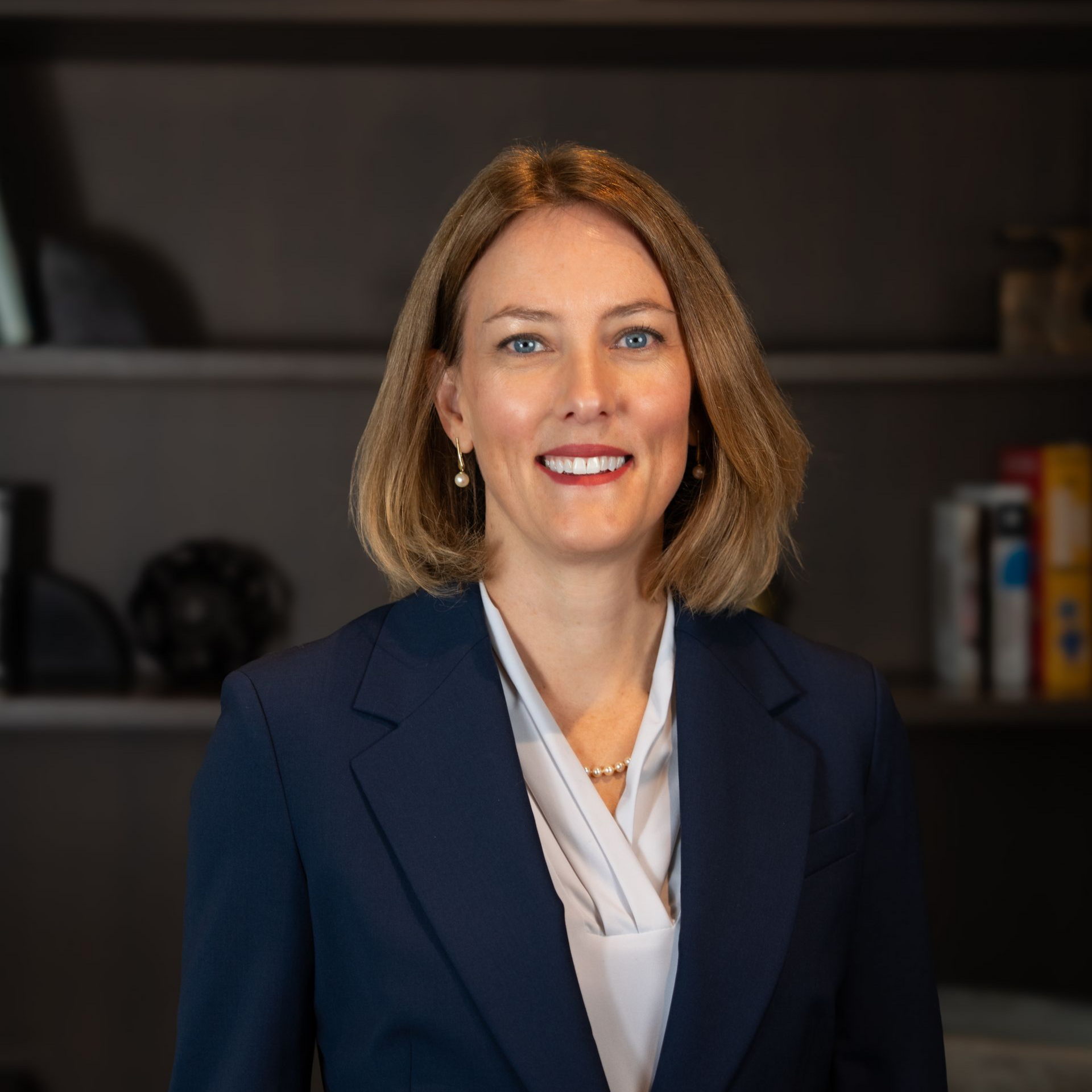 Professional headshot of a smiling woman in a navy blazer against a blurred office backdrop