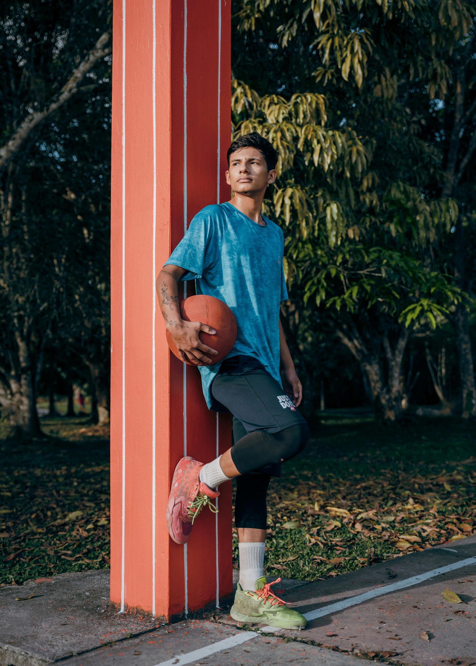 Persona con camisa azul apoyada en un pilar rojo, sosteniendo una pelota de baloncesto