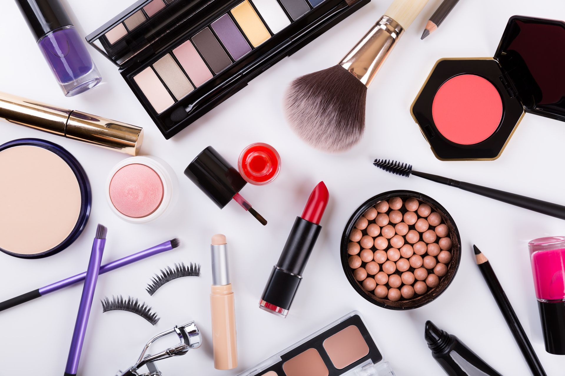 Makeup products scattered on a white surface, including eyeshadow, blush, lipstick, brushes, and mascara.