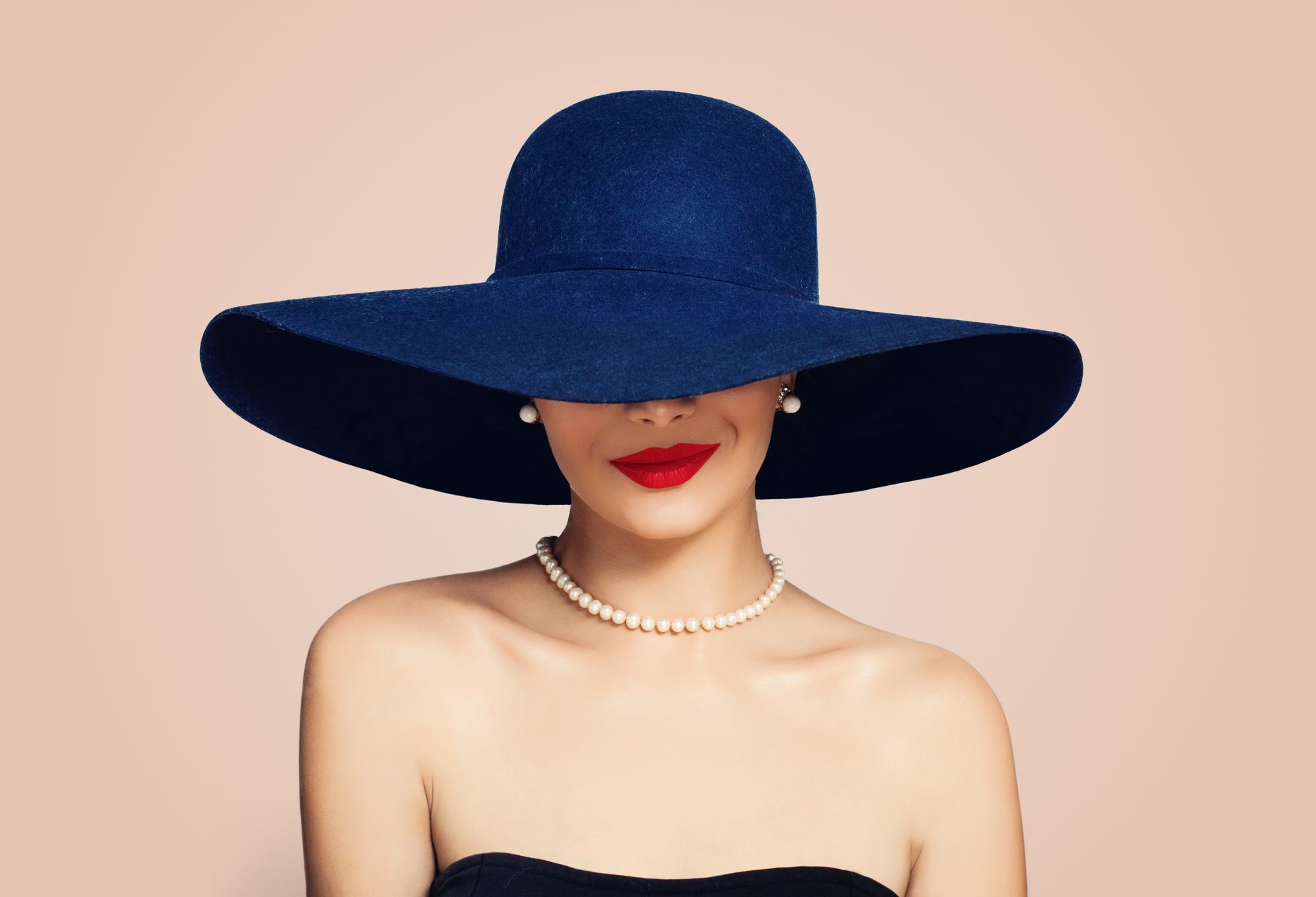 Woman in blue floppy hat, pearl necklace, red lipstick, against a pink background.