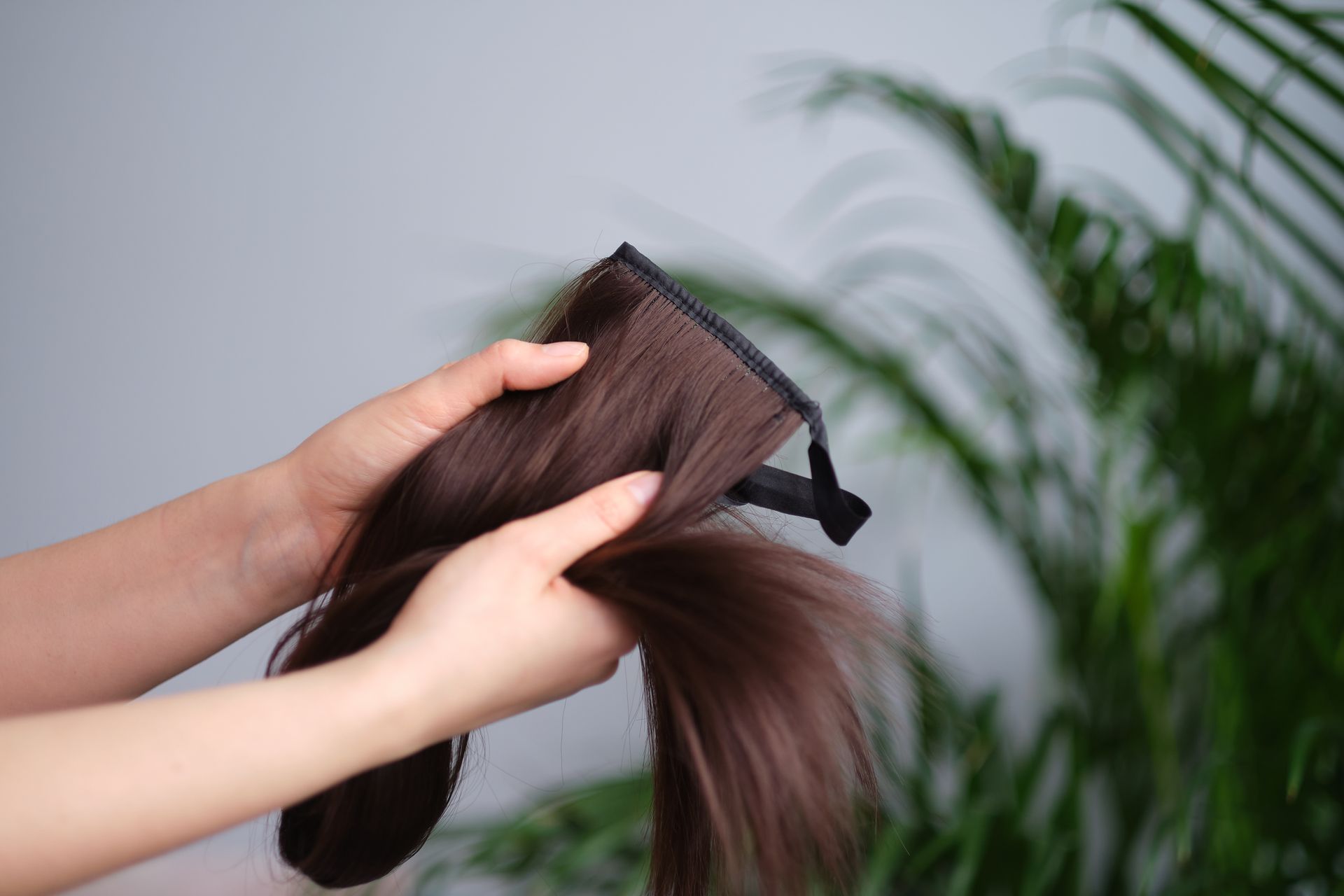 Hands holding a brown hair extension piece with a clip, against a blurry green plant background.