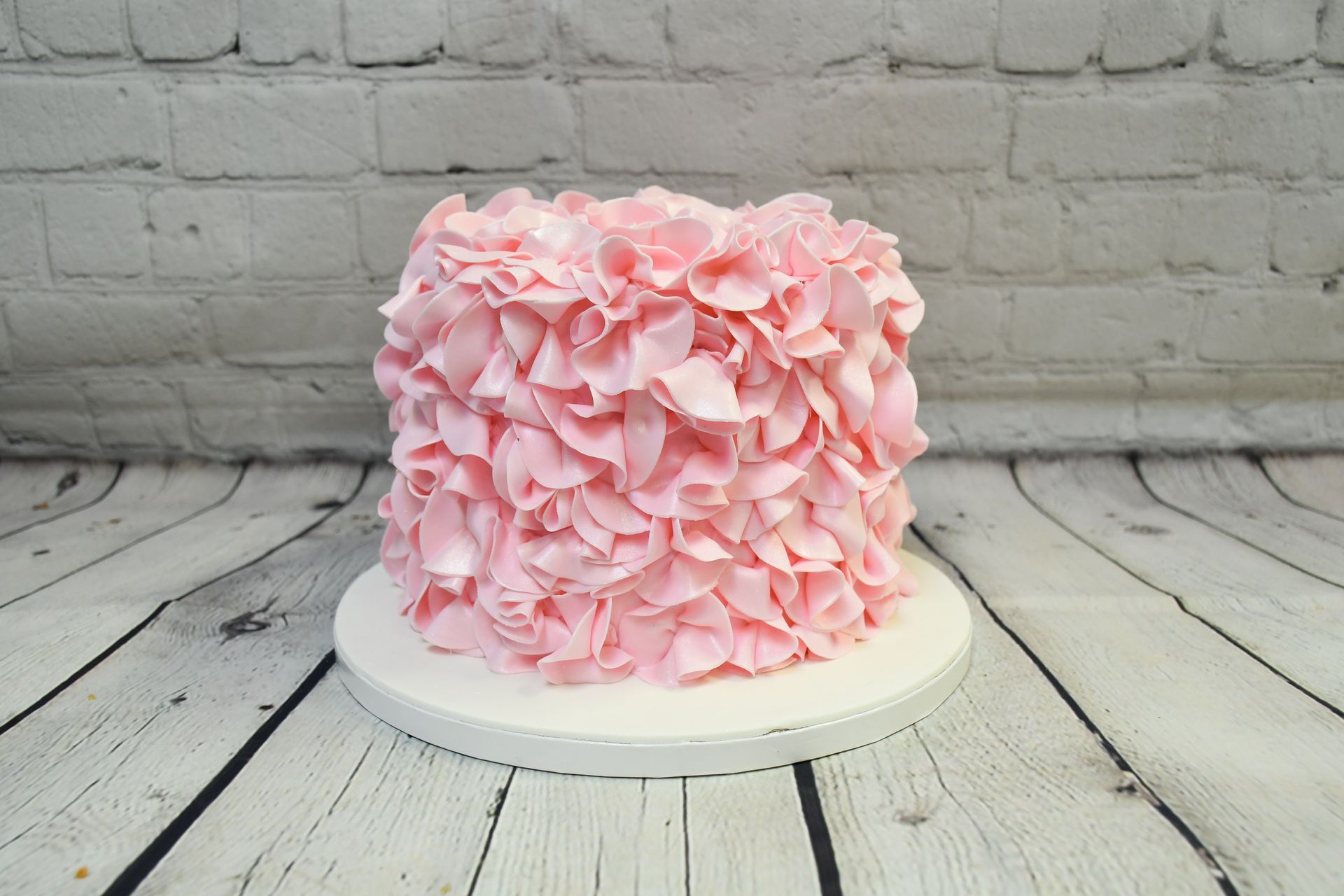A pink cake with ruffles on it is sitting on a white wooden table.