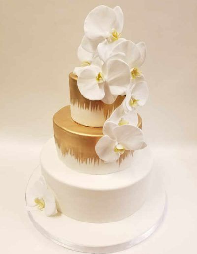 A white and gold wedding cake with white flowers on top.