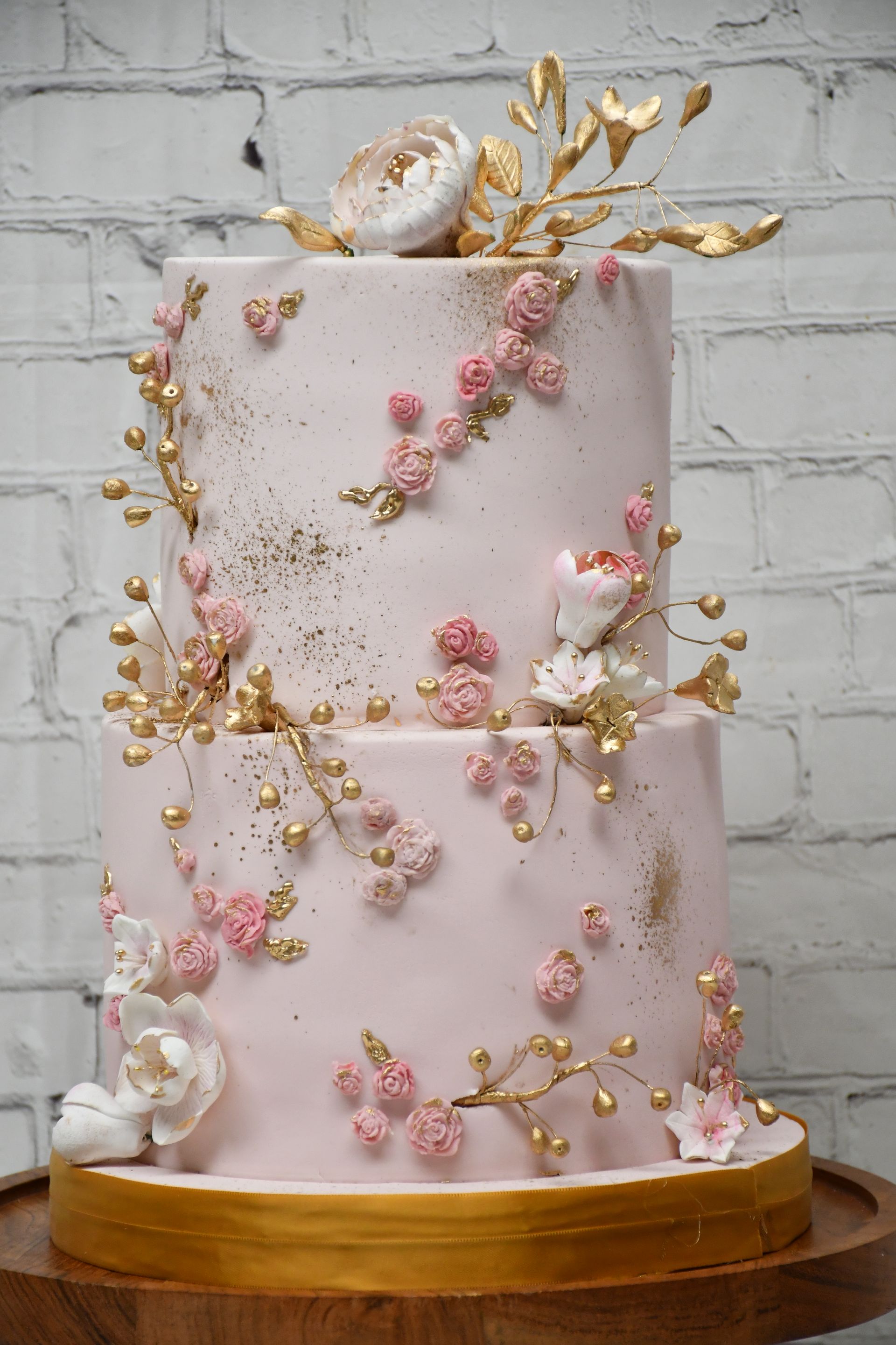 A pink and gold wedding cake with flowers and gold leaves on a wooden stand.