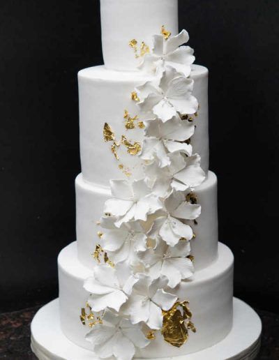 A white wedding cake with white flowers and gold leaf on it.