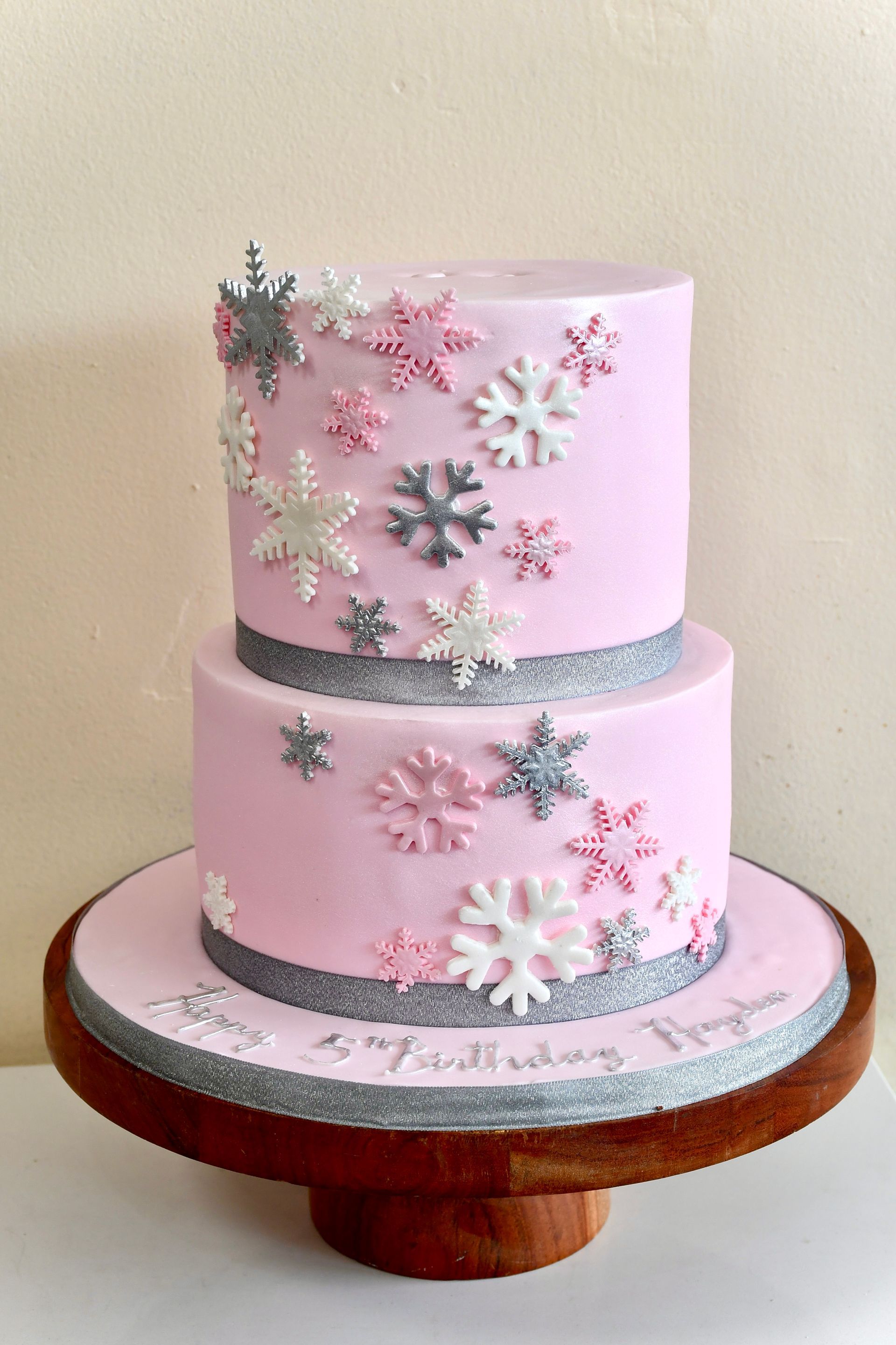 A pink cake with snowflakes on it is on a wooden cake stand.