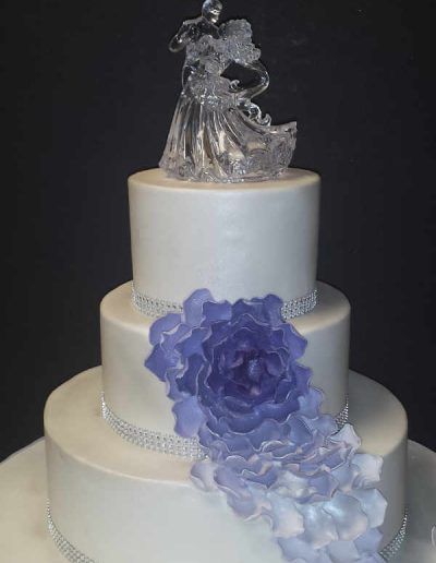 A white wedding cake with a purple flower and a bride and groom figurine on top.