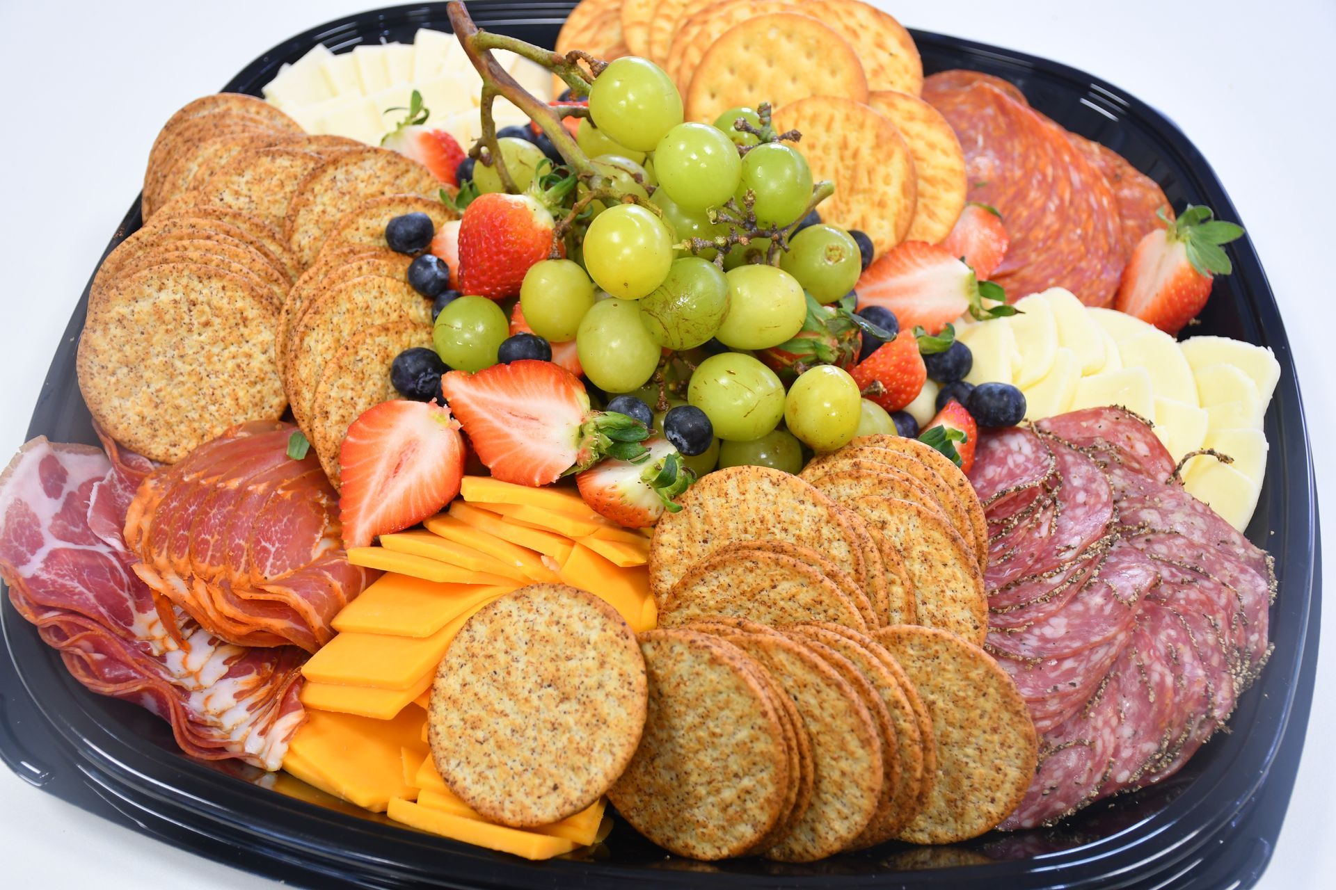 A tray of crackers , cheese , grapes , strawberries and meat on a table.