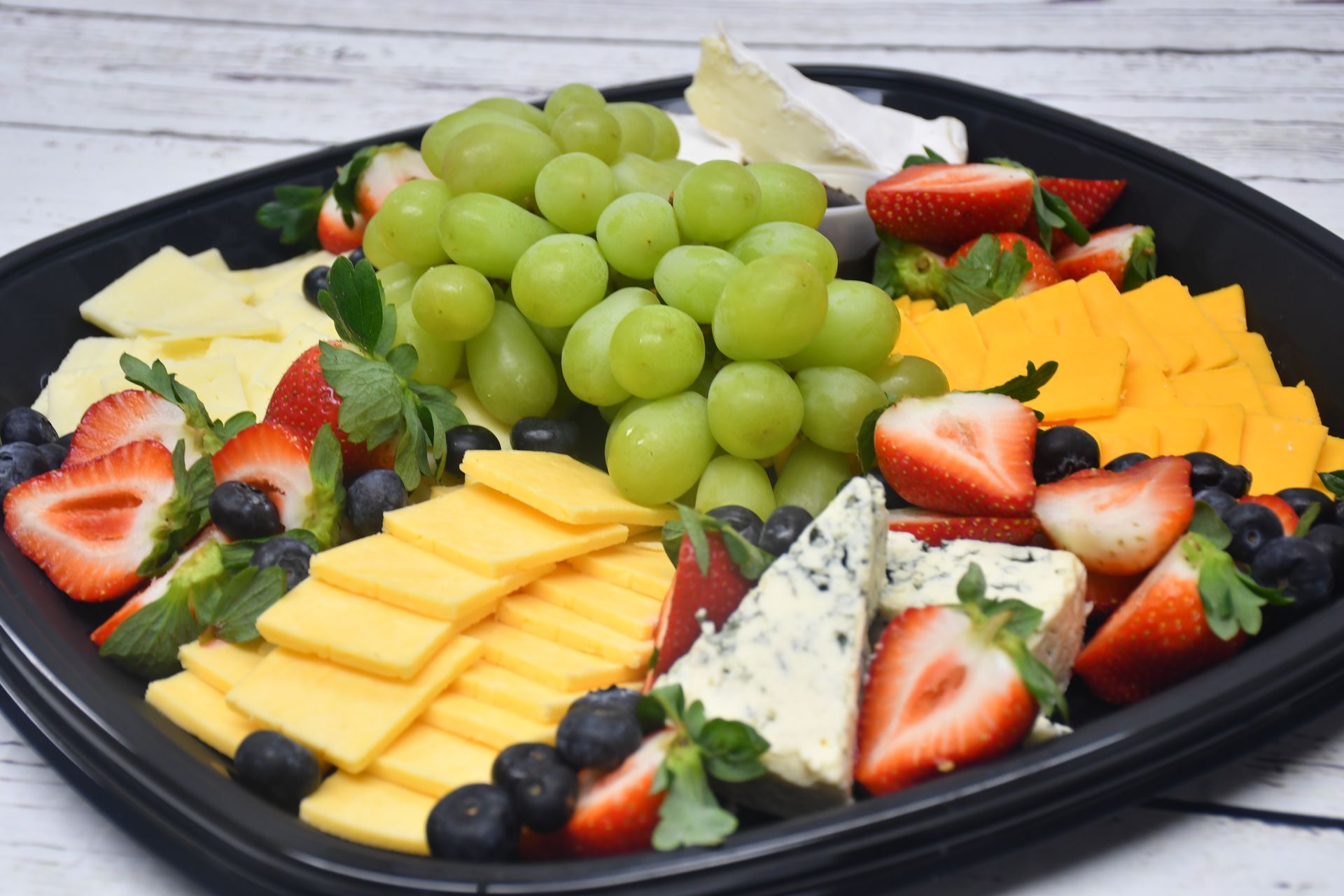 A black tray filled with cheese , grapes , strawberries and blueberries.