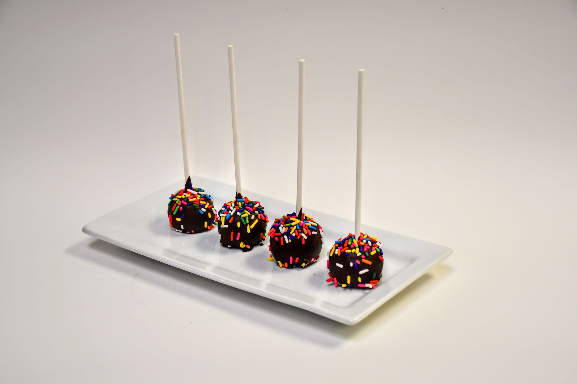 Four chocolate cake pops with sprinkles on a white plate.