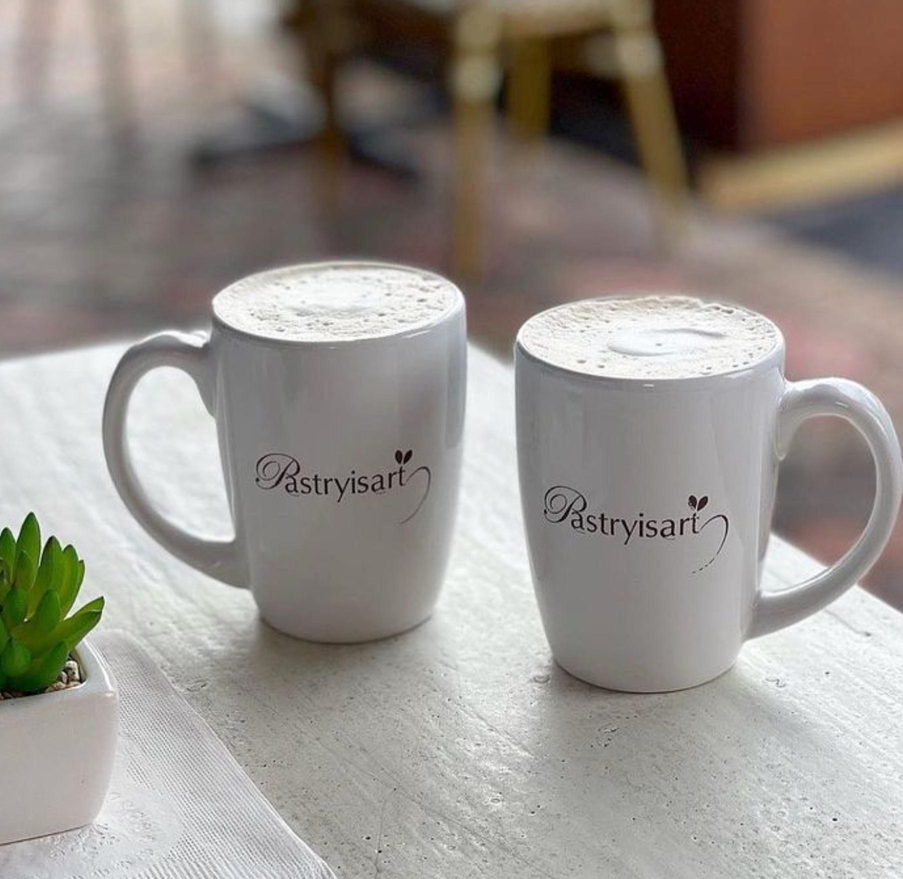 Two white coffee mugs with pastrysari written on them are sitting on a table.