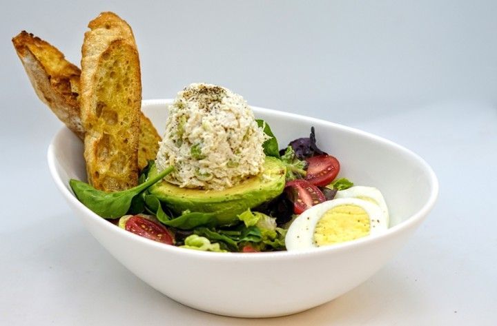 A salad with avocado , hard boiled eggs , tomatoes and toasted bread in a white bowl.
