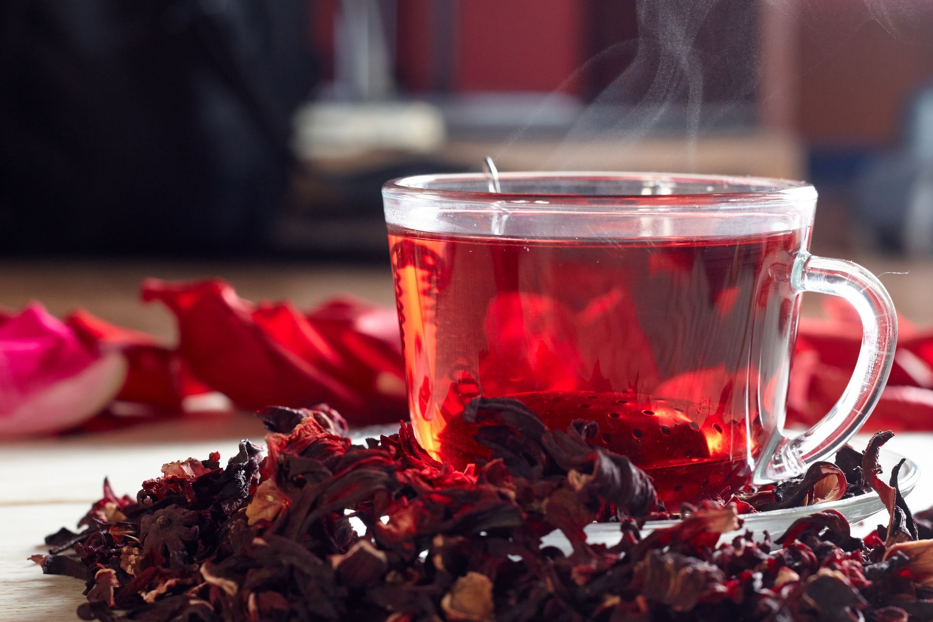 A cup of red tea is sitting on a saucer next to dried hibiscus flowers.