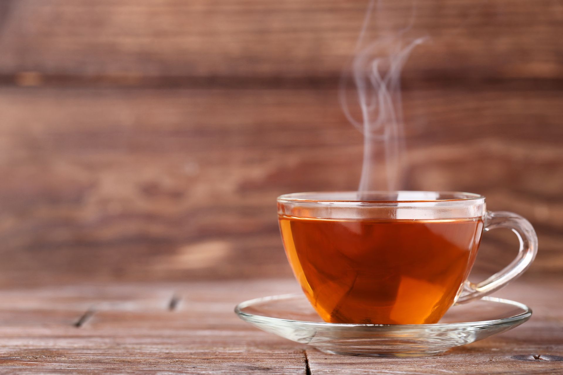 A cup of tea with steam coming out of it is on a saucer on a wooden table.
