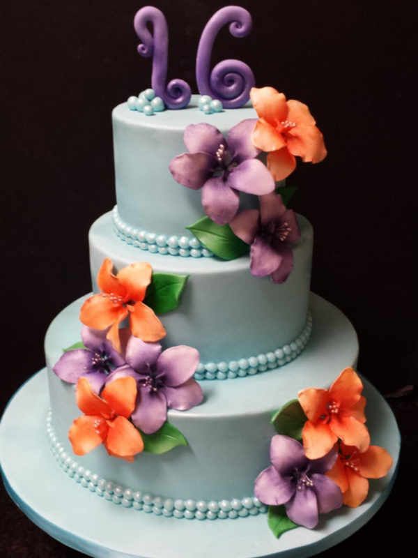 A blue cake with purple and orange flowers and the number 16 on top