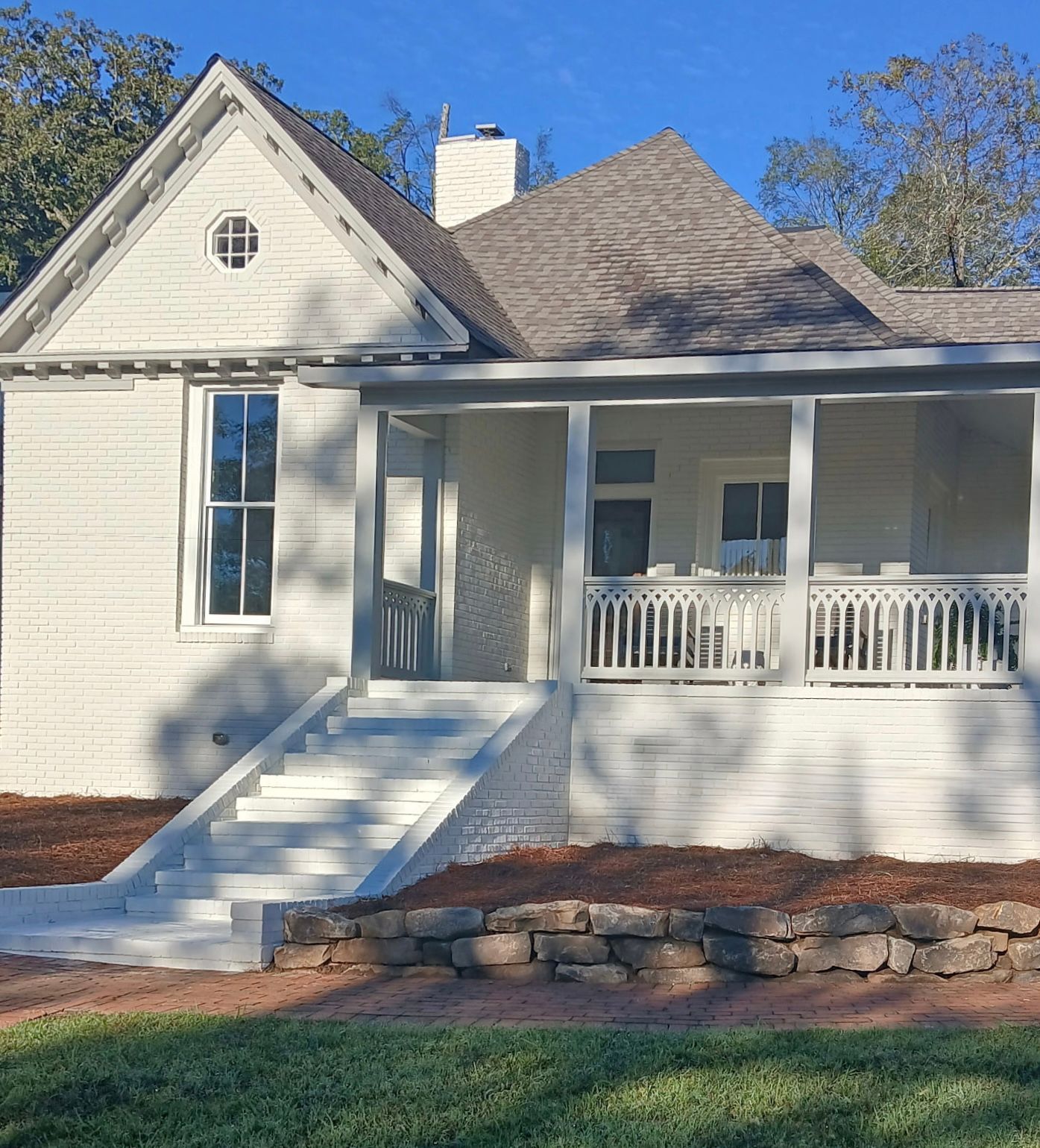 A single-story white house with a gable roof, front porch, and stone retaining wall on a sunny day.