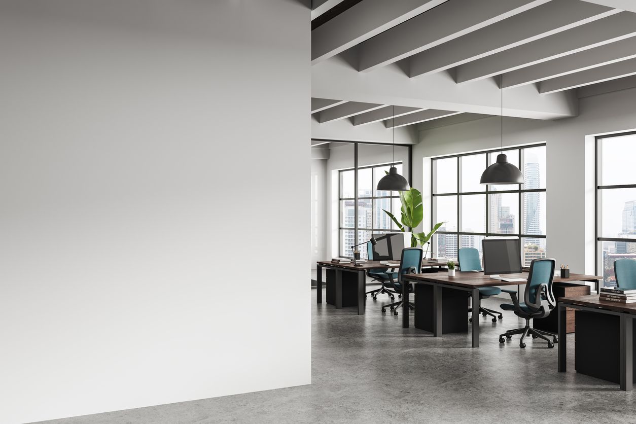 A modern, minimalist office space with an empty white wall in the foreground and rows of workstations with blue chairs.
