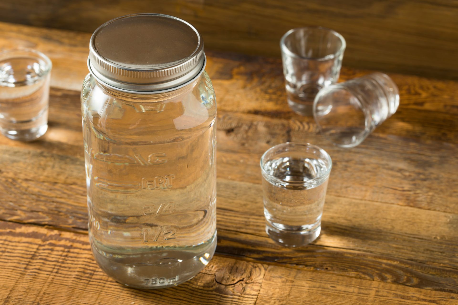 A mason jar filled with water and three shot glasses on a wooden table.