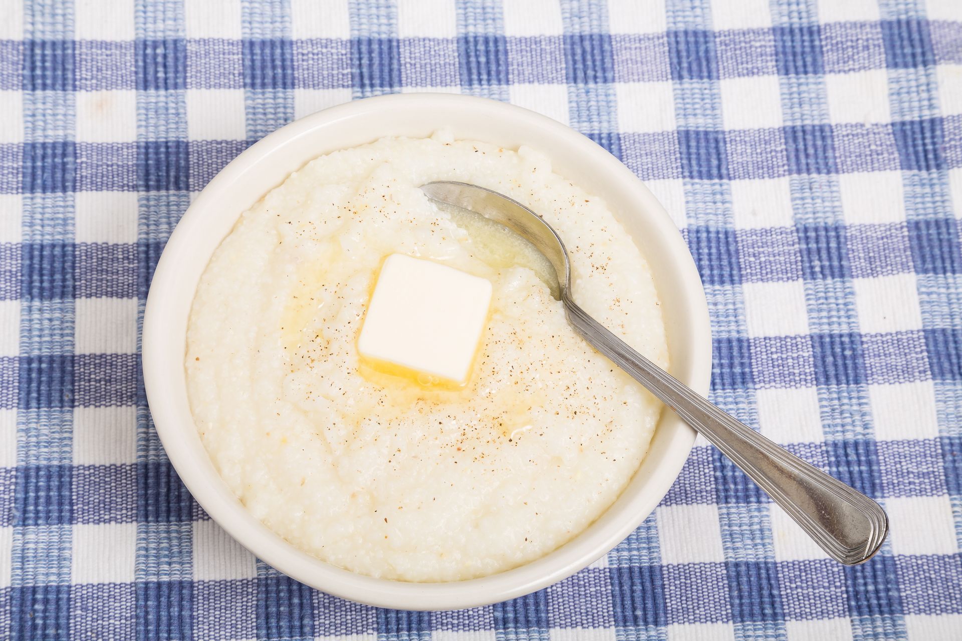 A bowl of grits with butter and a spoon on a checkered table cloth.