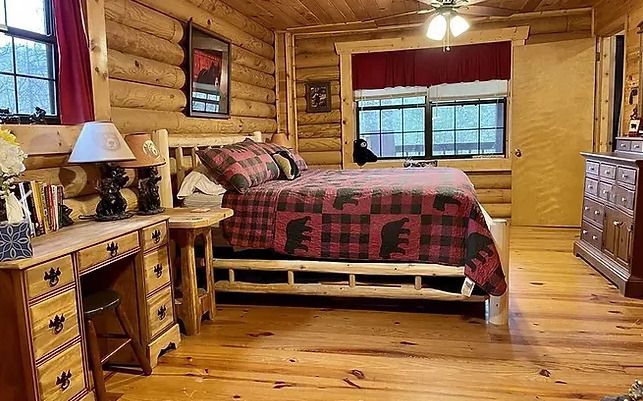 A bedroom in a log cabin with a bed , dresser , desk and window.