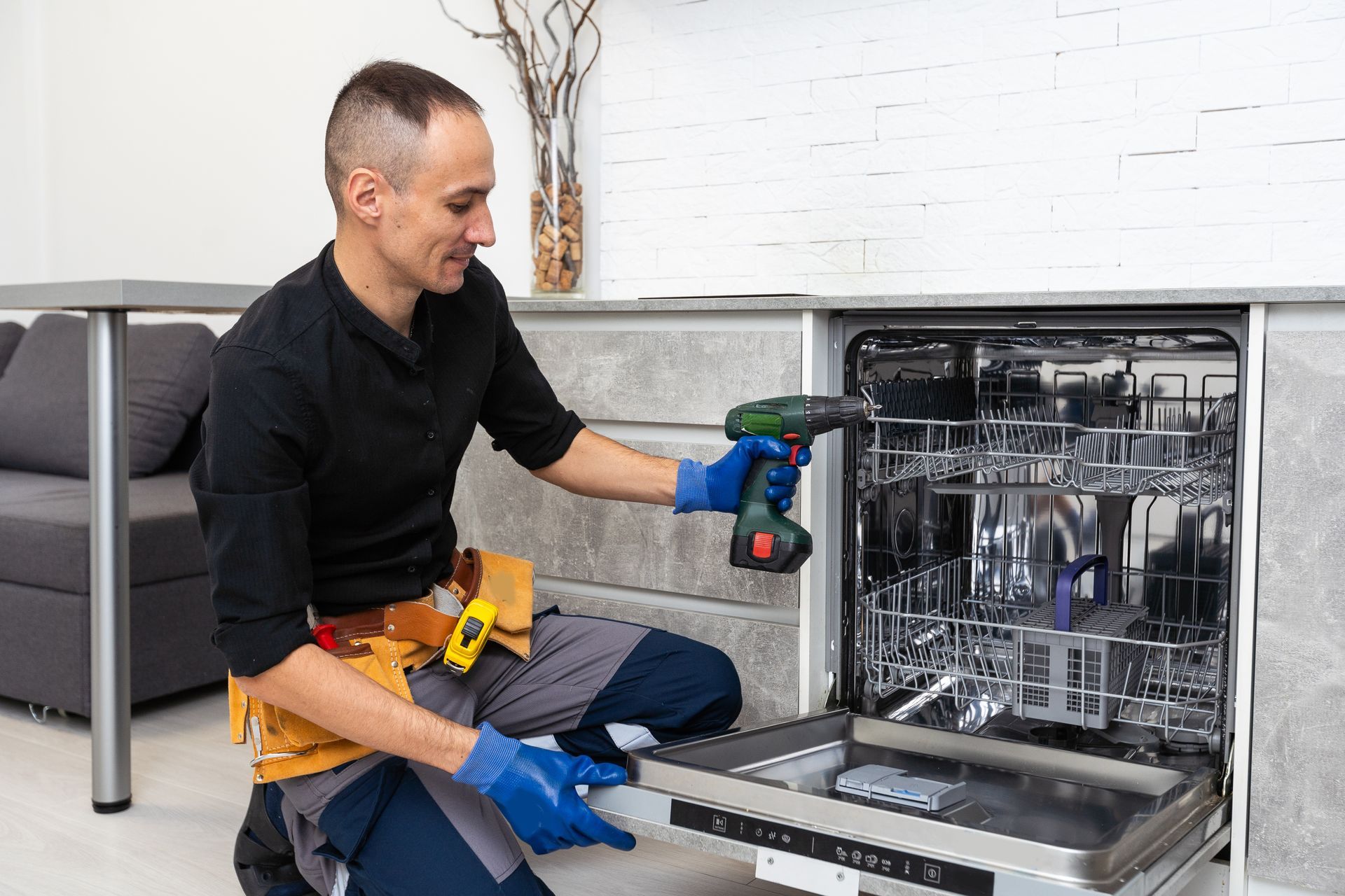 A man is fixing a dishwasher in a kitchen with a drill.