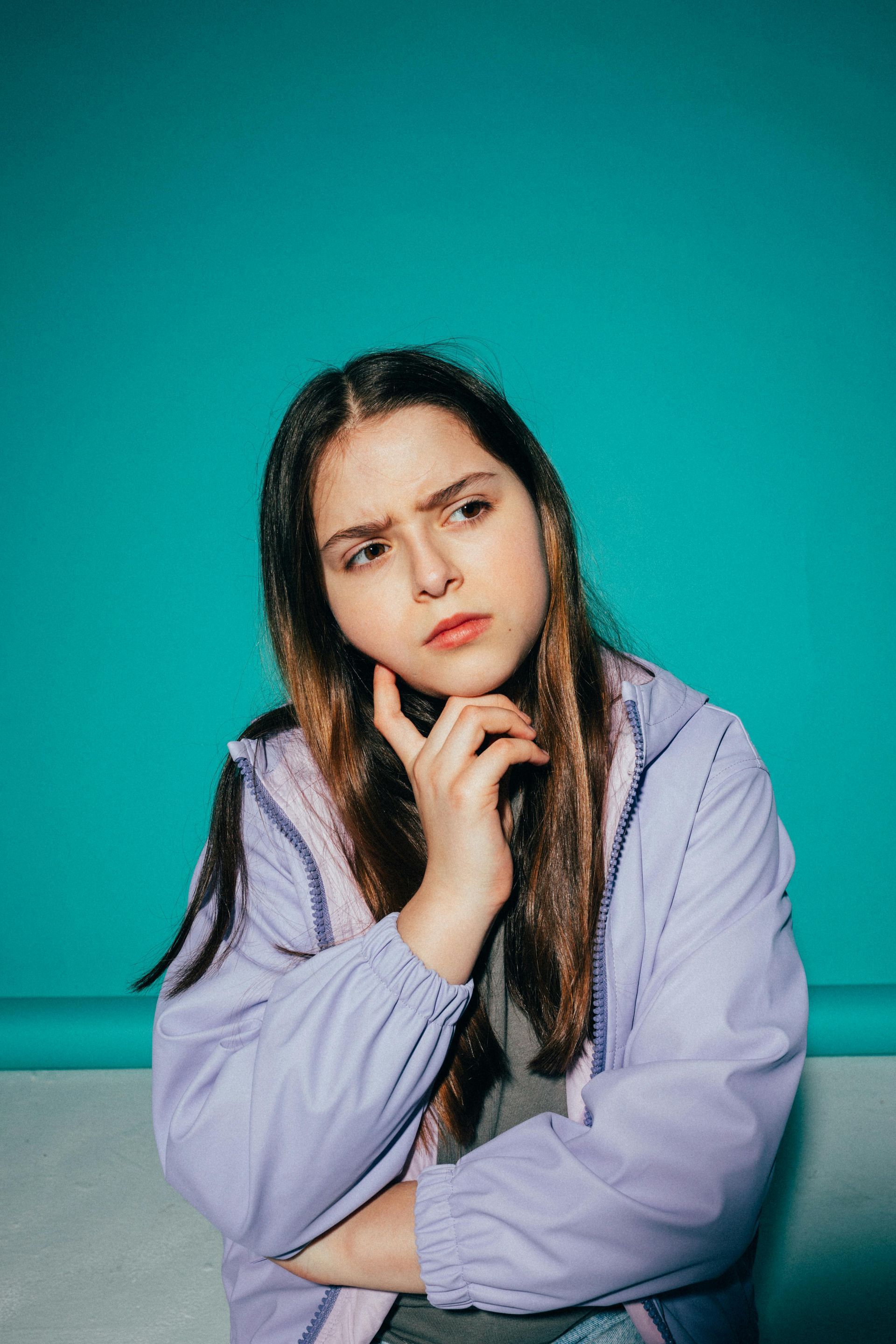 A young girl in a purple jacket is sitting on a couch with her hands on her chin.