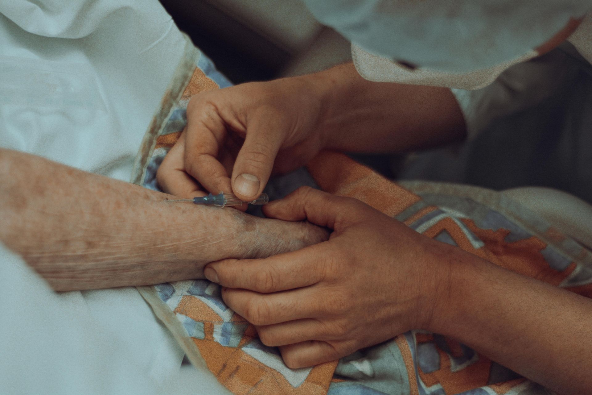Two hands gently holding an older person’s hand under a patterned blanket