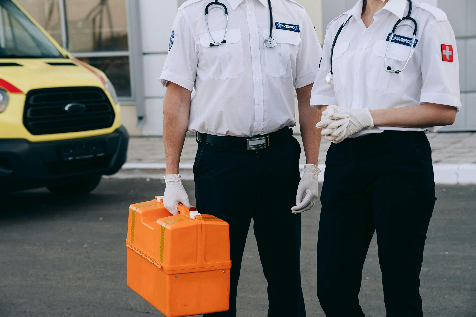 Two paramedics in white uniforms carrying an orange medical case beside an ambulance