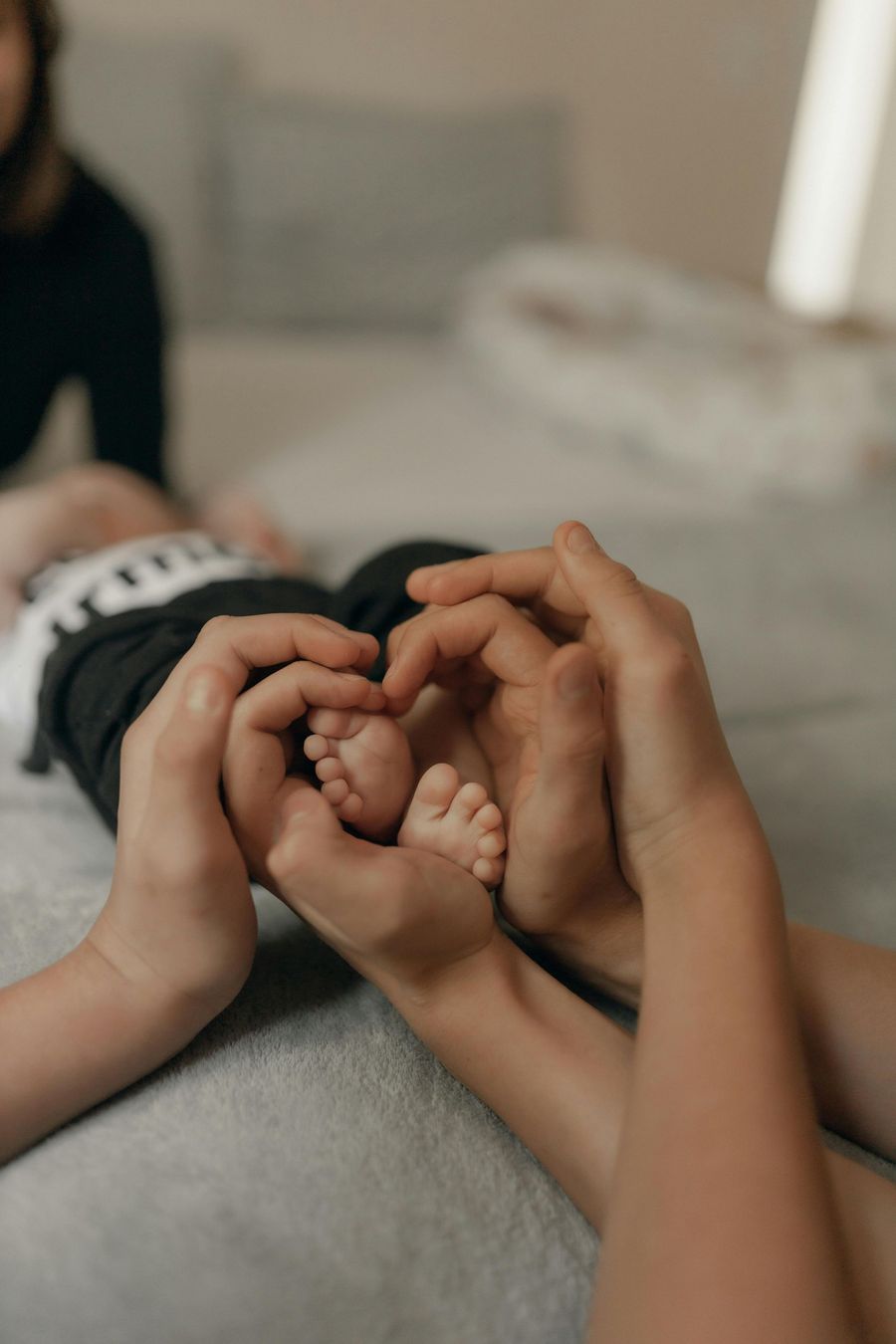Two adults hold a baby’s hands and feet on a couch, with a blurred living room in the background